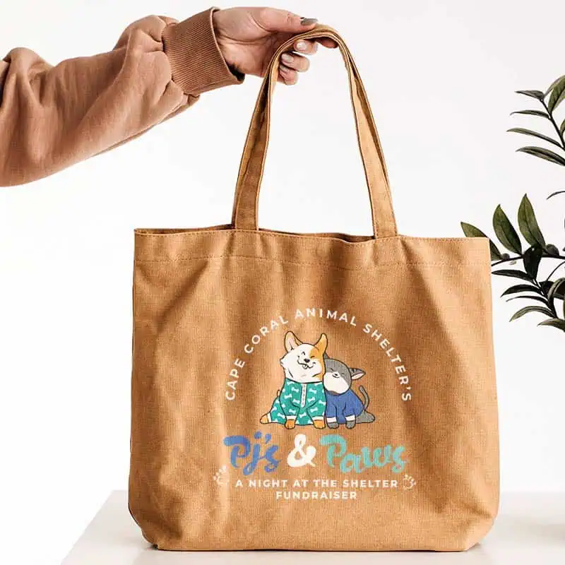 Young woman holding up a Cape Coral Animal Shelter PJs & Paws Fundraiser light brown canvas tote bag