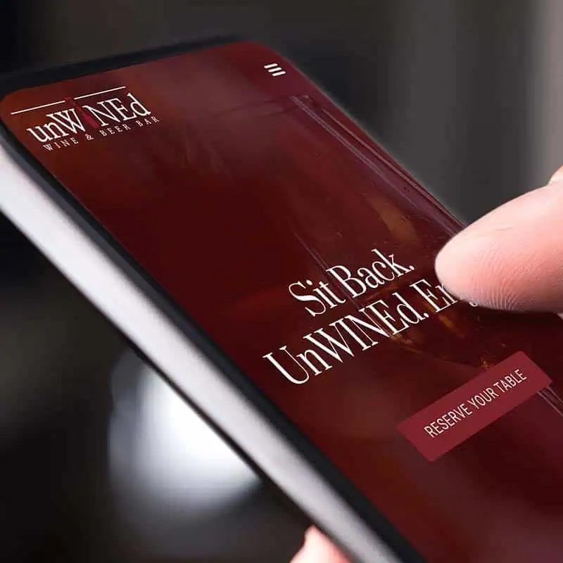 Close-up view of person holding mobile phone displaying UnWINEd Wine+Beer Bar responsive website